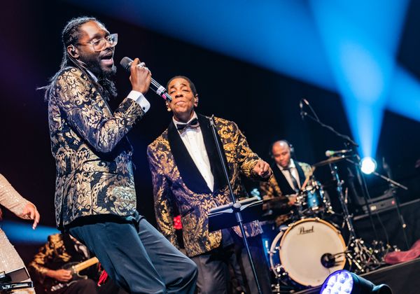 Dvaja speváci v zlatých brokátových sakách spievajú naživo na pódiu, za nimi bubeník